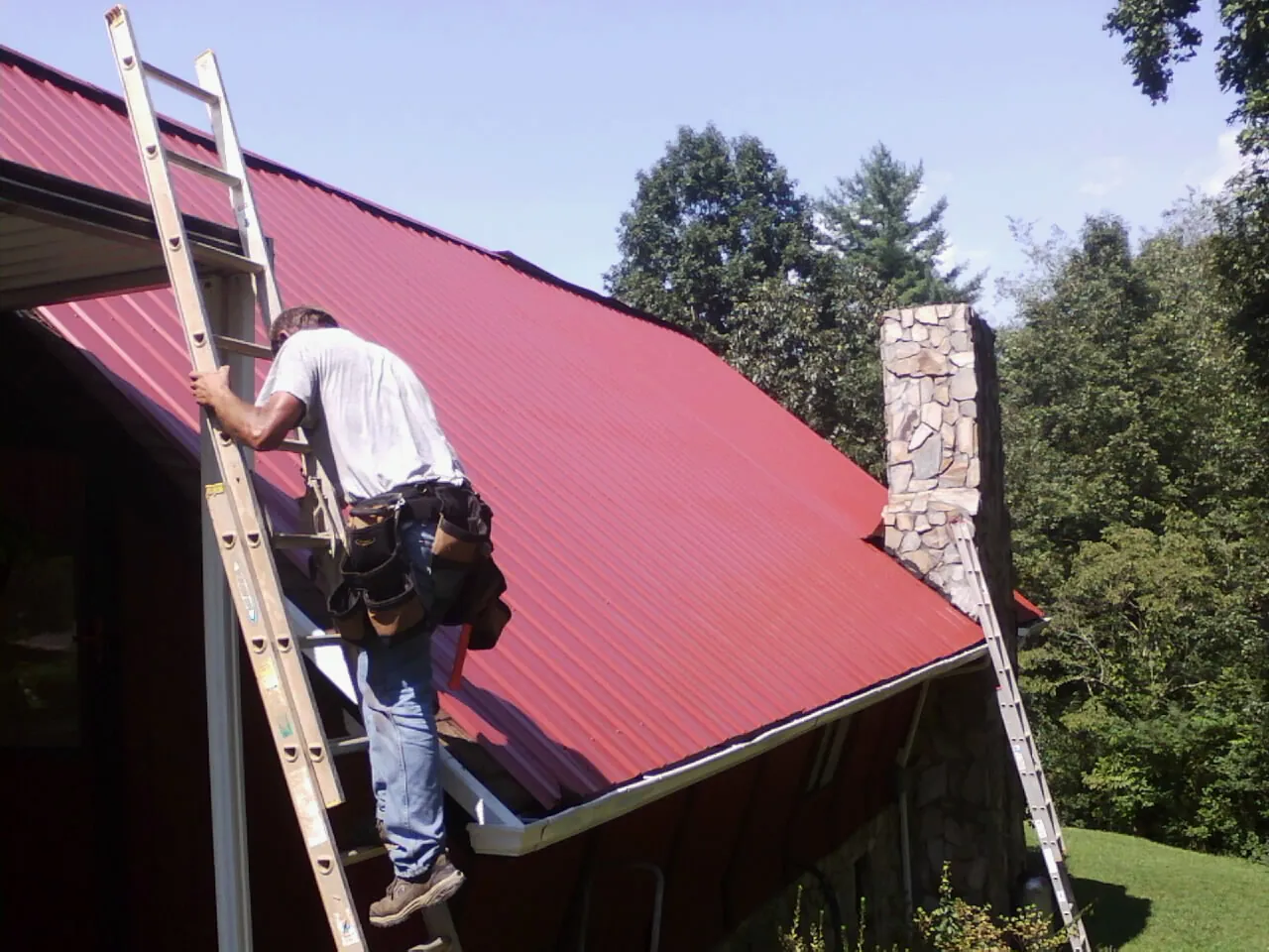 Roofer on a ladder beside a red metal roof and stone chimney — Soffit & Fascia Repair by Roofing Brothers in Imnaha